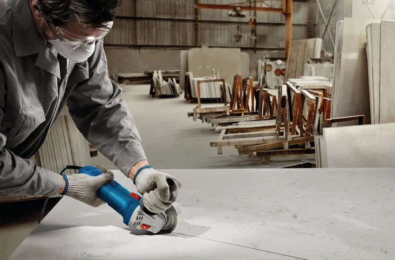 A person wearing safety equipment uses a grinder to cut a marble slab in a workshop.