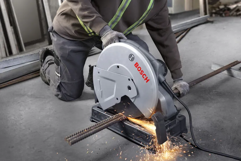 A person wearing safety equipment cuts metal rebar with a chop saw, creating sparks.