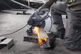 A person wearing safety equipment cuts metal tubing with a chop saw, sparks flying.