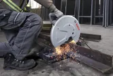 A person wearing safety equipment operates a chop saw cutting a steel beam.