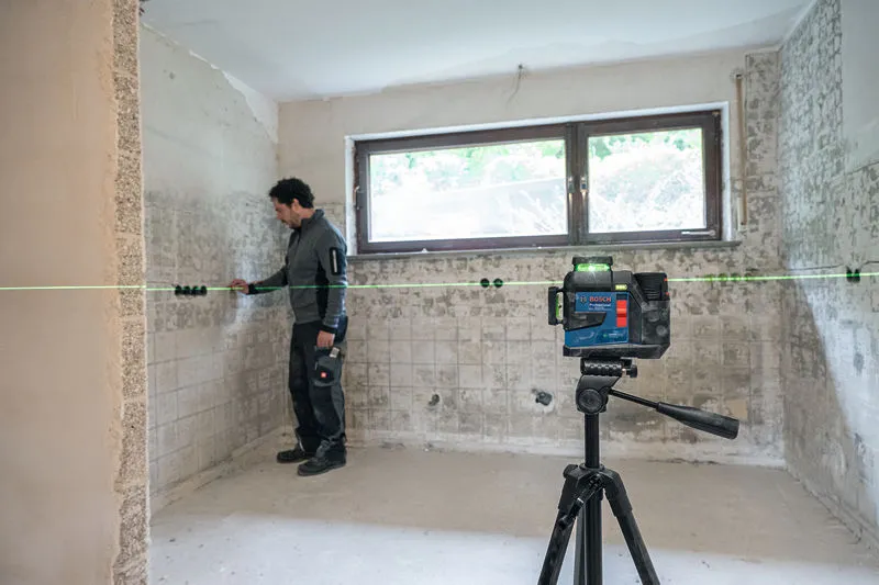 A worker wearing safety equipment aligns tiles using a laser leveling tool on a tripod.