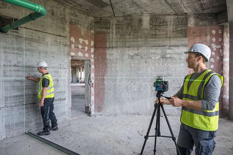 Two workers wearing safety equipment use a laser leveling tool to mark reference lines on a wall.