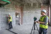 Two workers wearing safety equipment use a laser leveling tool to mark reference lines on a wall.