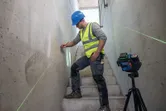A person wearing safety equipment marks a wall using a laser leveling tool on a tripod.