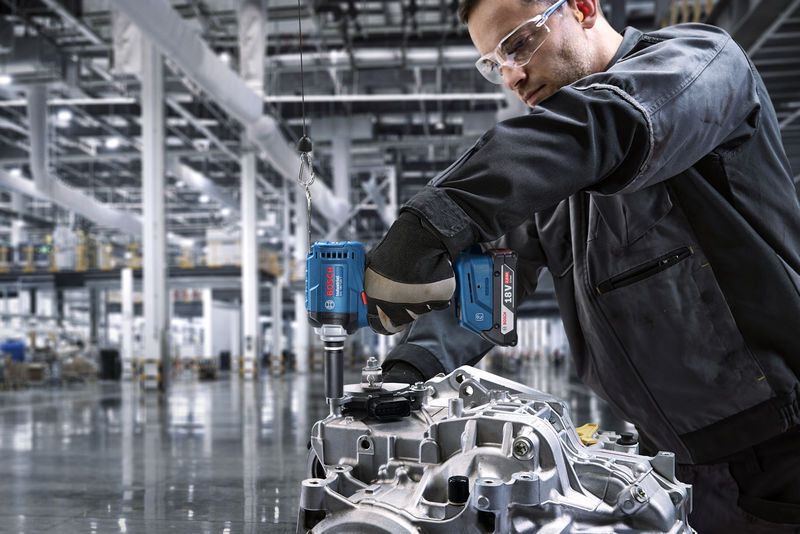 A person wearing safety equipment uses a cordless drill on an engine part.
