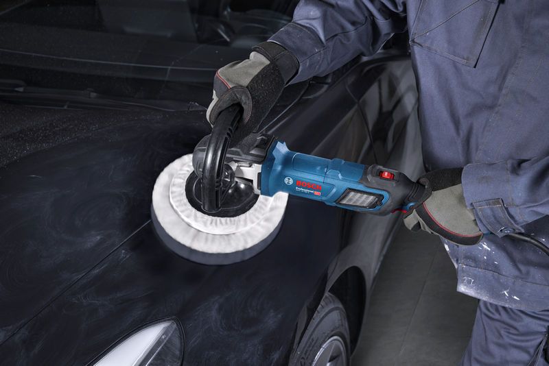 A person wearing safety equipment uses a polisher on a black car hood.
