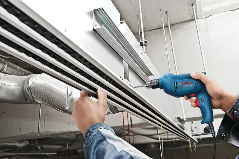 A person uses a power drill to bore a hole into overhead metal framing.