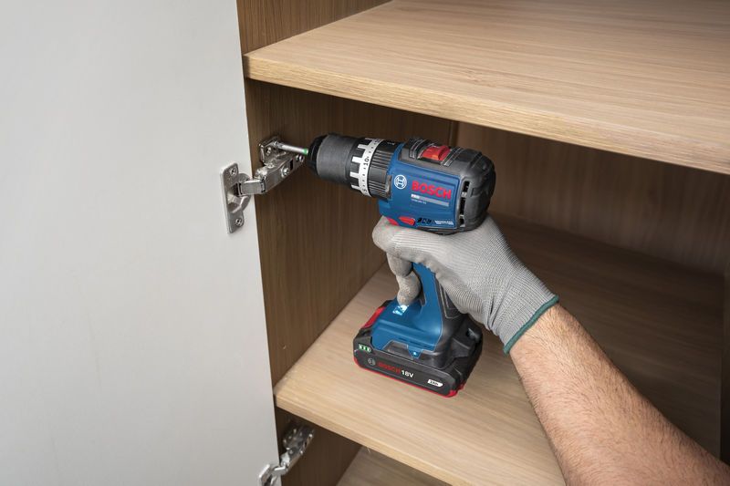 A person wearing safety equipment uses a cordless drill to install a cabinet hinge.