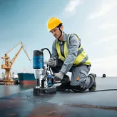 A person wearing safety equipment operates a magnetic core drill on a steel surface.