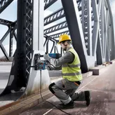 A person wearing safety equipment operates a magnetic core drill on a steel bridge.