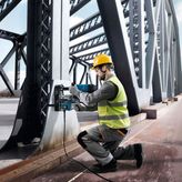 A person wearing safety equipment uses a magnetic core drill on a steel bridge beam.
