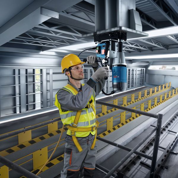 A person wearing safety equipment uses a magnetic core drill on a steel beam.