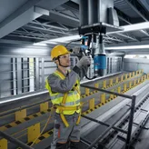 A person wearing safety equipment uses a magnetic core drill on a steel beam.