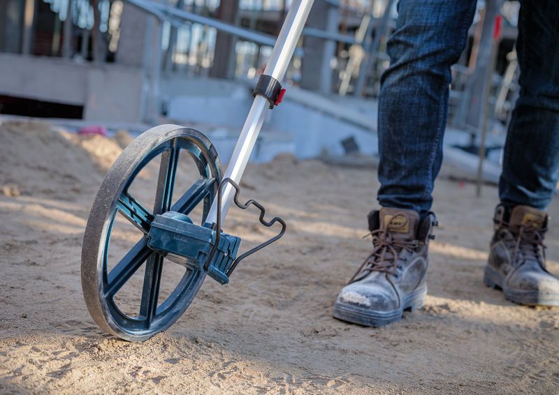 A person in work boots uses a measuring wheel on a sandy construction site.