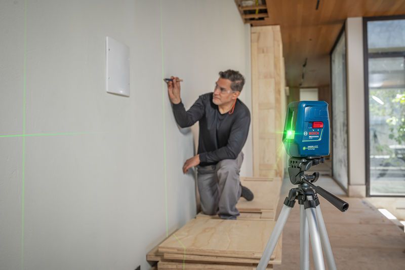 Person marks a wall using a laser leveling tool projecting green lines.