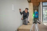 Person marks a wall using a laser leveling tool projecting green lines.