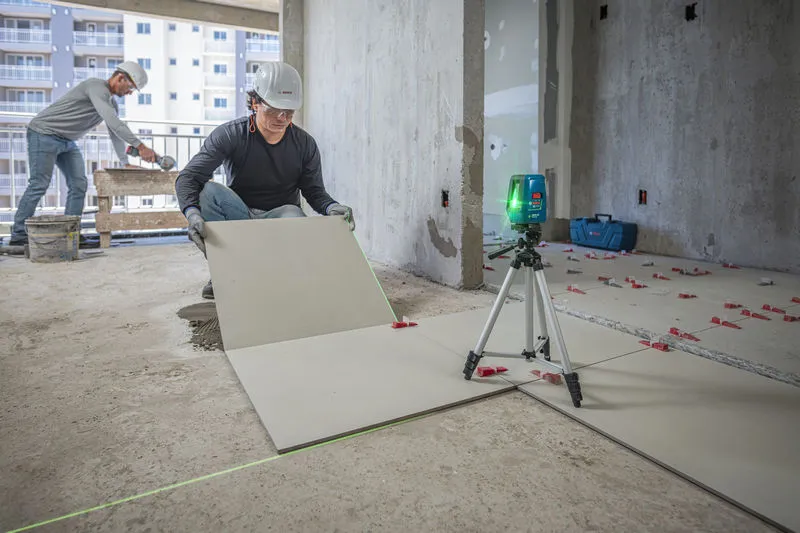 A person wearing safety equipment aligns floor tiles using a laser leveling tool.