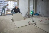 A person wearing safety equipment aligns floor tiles using a laser leveling tool.