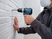 A person wearing safety equipment drills into a brick wall with a cordless impact drill.