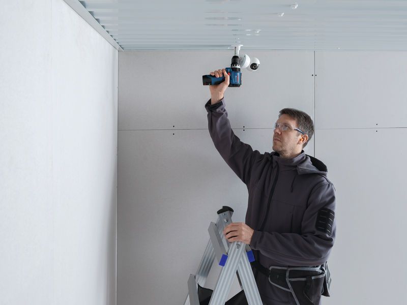 A person wearing safety equipment uses a cordless drill on a ceiling while standing on a ladder.