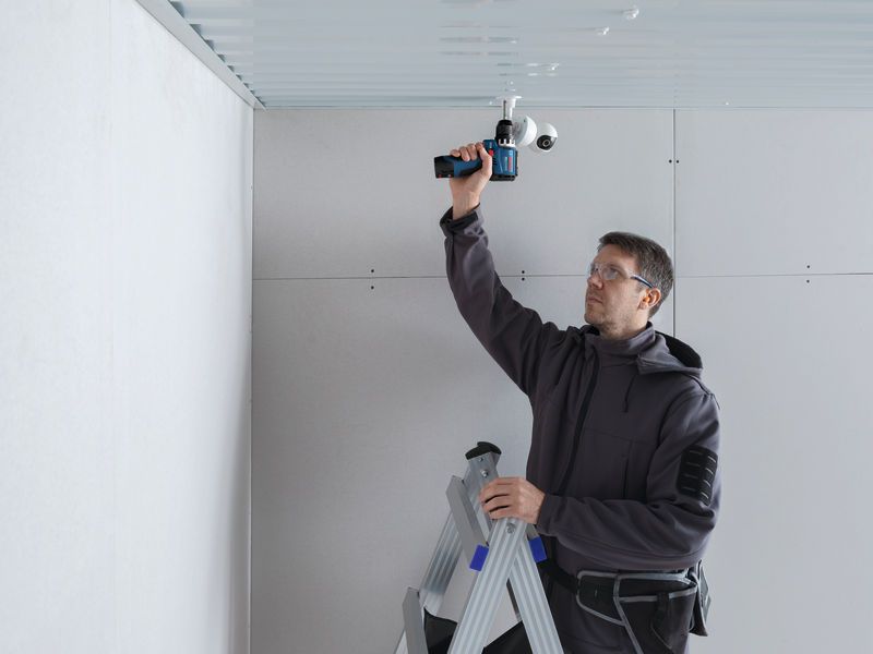 A person wearing safety equipment installs a security camera on a ceiling using a cordless drill.