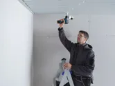 A person wearing safety equipment installs a security camera on a ceiling using a cordless drill.
