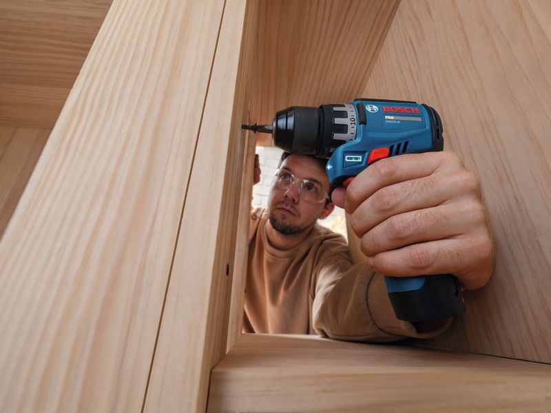 A person wearing safety equipment uses a cordless drill driver on a wooden frame.
