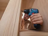 A person wearing safety equipment uses a cordless drill driver on a wooden frame.