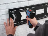 A person secures a metal bracket to a white brick wall with a cordless drill.