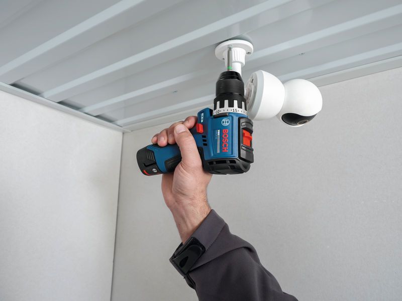 A person uses a cordless drill to install a security camera on a ceiling.