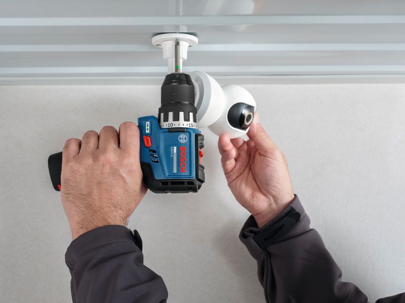 A person installs a security camera on a ceiling using a cordless drill.