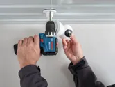 A person installs a security camera on a ceiling using a cordless drill.