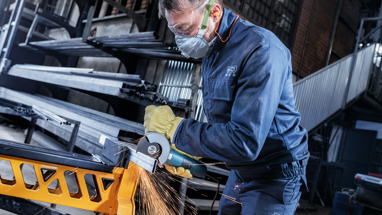 Person wearing safety equipment grinds a metal beam with an angle grinder.