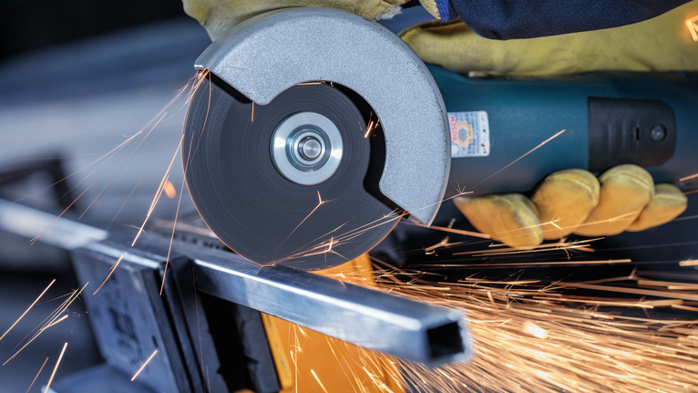 Person wearing safety equipment cuts metal with an angle grinder, creating sparks.
