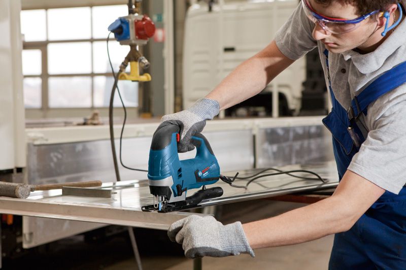 A person wearing safety equipment uses a jigsaw to cut a metal sheet.