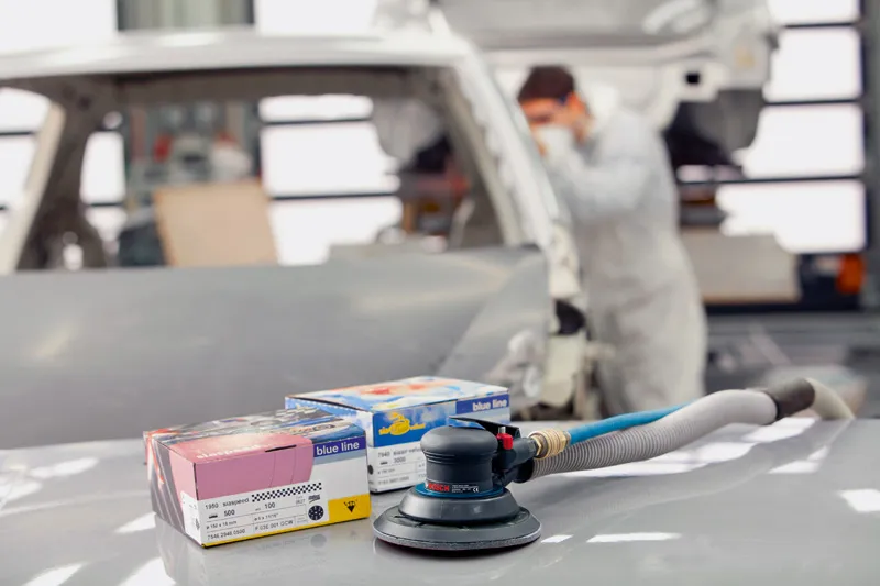 A person wearing safety equipment works near a pneumatic sander and sanding discs in an auto shop.