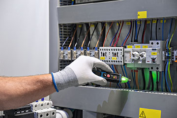 A hand wearing a white glove holds a black multimeter near electrical connections in a control panel. Wires in various colors are visible, along with labels and warning signs, indicating a professional electrical environment.