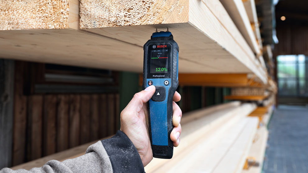 A hand holding a Bosch Professional moisture meter displays a reading of 12.0% near stacked wooden beams.