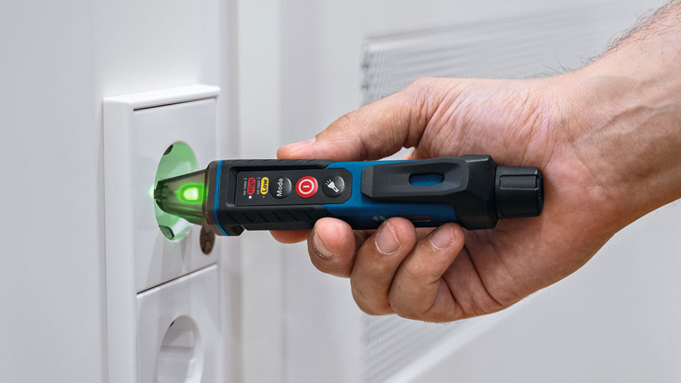 A Bosch Professional voltage tester is held by a hand, with a green light indicating functionality, near an electrical outlet.